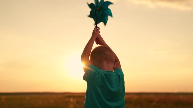 Child childhood. Family outing, nature. Kid boy runs through summer field holding toy pinwheel, sunny day. Happy child plays with toy pinwheel outdoors. Child nature. Child son, dream in park sunset