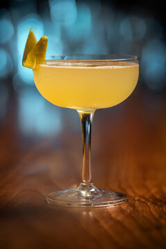 Elegant cocktail, the Bees Knees in coupe glass. Lemon twist adorns the rim. Warm wood table contrasts cool background. Soft bokeh lights enhance drink's allure. Perfect for bar, lounge