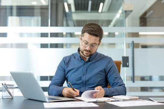 Professional businessman working on financial report, focused entrepreneur analyzing business documents and laptop data in modern corporate office