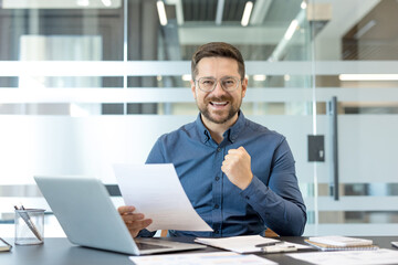 Obraz premium Success and career achievement, happy businessman celebrating a project milestone with paperwork and laptop in a modern office