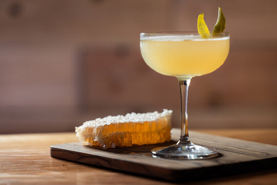 Elegant cocktail, the Bees Knees in coupe glass. Lemon twist adorns the rim. Warm wood table and honey comb garnish.