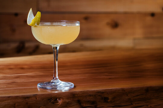 Elegant cocktail, the Bees Knees in coupe glass. Lemon twist adorns the rim. Warm wood table. Perfect for bar, lounge