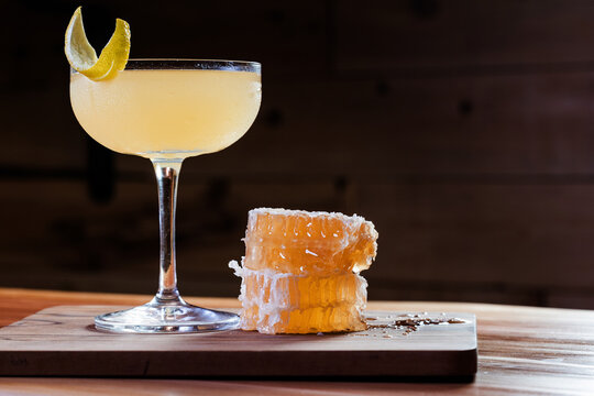 Elegant cocktail, the Bees Knees in coupe glass. Lemon twist adorns the rim. Warm wood table and honey comb garnish.