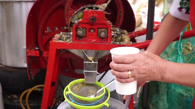 Vendor pouring freshly pressed sugarcane juice with a ladle into a paper cup next to a red extraction machine