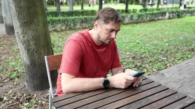 Side view of Caucasian bearded man in his 40s using smartphone at wooden table on outdoor cafe terrace in park in daylight