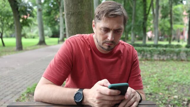 Caucasian bearded man in his 40s wearing red T-shirt using smartphone at wooden table with park in background in daylight
