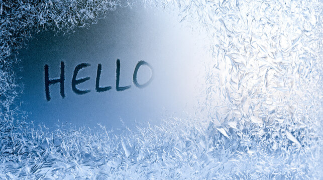 The word hello is written on a condensated frozen window glass framed with beautiful ice frost patterns. Cold winter morning message concept on a misty glass closeup