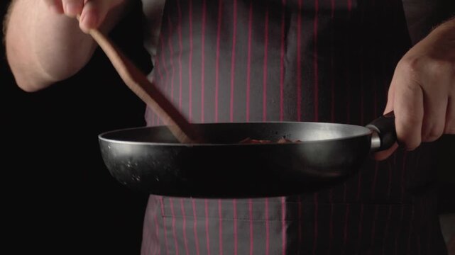 Chef in striped apron flips pancake with wooden spatula in black frying pan, showcasing cooking technique and precision in a dimly lit kitchen environment with dark background