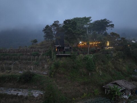 Kecamatan Getasan, Jawa Tengah, Indonesia - 08 September 2026: Aerial view of Merbabu View Cafe on a hillside with terraced fields, warm glowing lights, and a misty mountain backdrop.