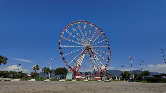 Panoramic view of a large red and white Ferris wheel slowly rotating in an amusement park on a sunny day, with a beautiful clear blue sky and mountains in the background in Batumi Georgia