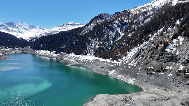 turquoise reservoir bends between steep snowcapped slopes rugged rock face descends toward shoreline forested mountainside frames lai da marmorera aerial drone footage graubunden switzerland