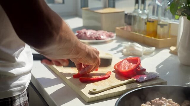 Person slices red bell pepper with knife on wooden cutting board. Preparing fresh vegetables for cooking and meal preparation