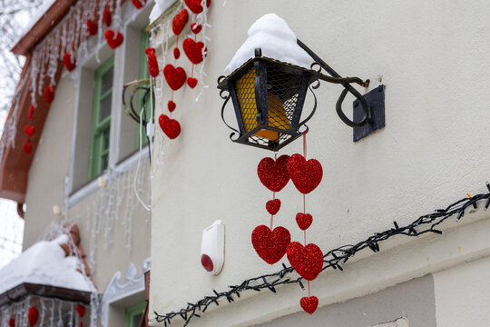 Red heart decorations hanging from outdoor wall lantern on snowy building for Valentine&rsquo;s Day