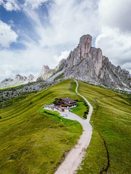 San Vito di Cadore, Italy - 21 August 2024: Aerial view of Giau Pass with a mountain hut in green meadows and a winding path leading towards rocky peaks under a cloudy sky.