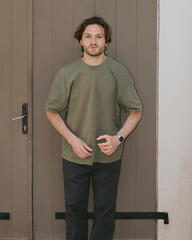 A young man in an oversized olive-colored T-shirt and dark trousers stands on the street by an old wooden door. © Vika