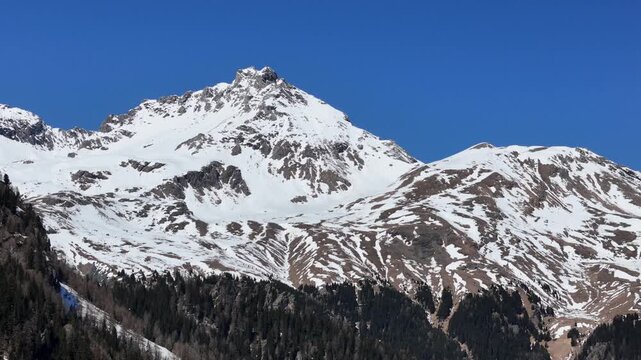 craggy snowcapped summit rises above patchy scree ridge steep rock face drops toward forested slopes descending julier pass mountain pass aerial drone footage graubunden switzerland clear blue sky