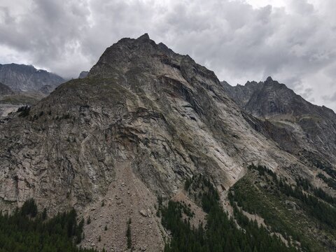 Aerial view of a rugged mountain peak with steep rocky slopes and a dense coniferous forest at the base under a cloudy sky in Italy.