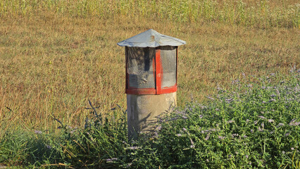 A weathered concrete ventilation pipe with a wire mesh cage stands among wild mint plants in a dry grassy field © hecos