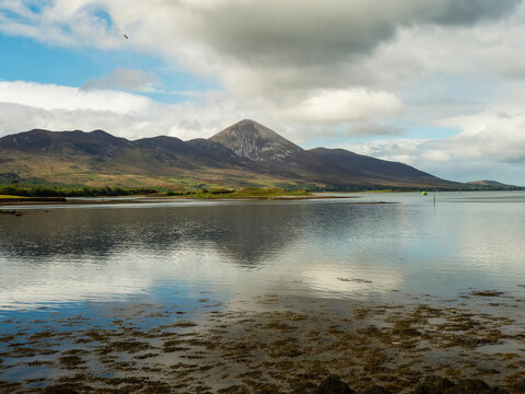Coastal landscape with rocky shore and distant mountains under cloudy sky. Still waters reflect overcast tones. Scene feels calm and isolated. View on Croagh Patrick, county Mayo, Ireland.