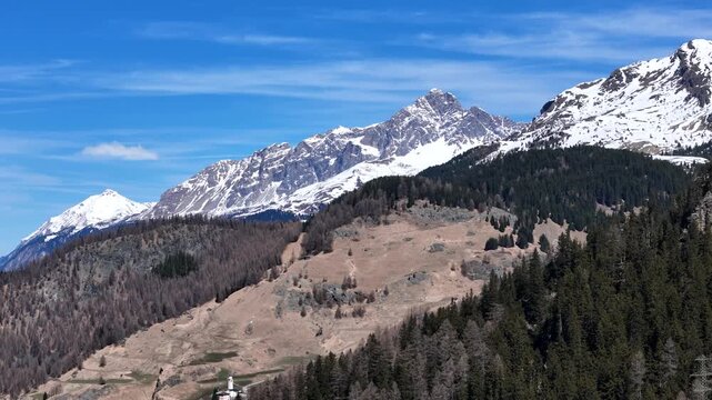forested mountainside climbs toward snowcapped sawtooth ridge scattered hamlet rooftops dot distant valley floor below meadow clearings near julier pass aerial drone footage graubunden switzerland
