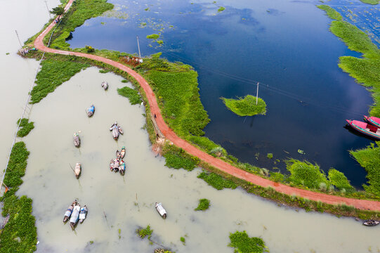 Aerial view of traditional wooden boats floating on the calm waters of a river, separated by a narrow dirt path from a deeper blue wetland area in Shariatpur, Dhaka Division, Bangladesh.