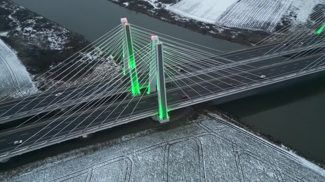 Aerial view captures cars driving along highway through the illuminated bridge at evening. Autos riding at speed road at cold season. Cable-stayed bridge spanning a river and snow-covered terrain