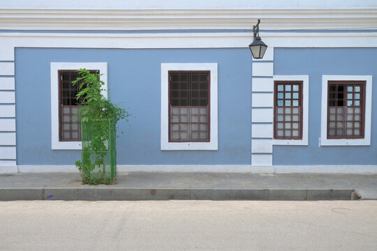 Straight-on architectural wall background featuring a light pastel blue facade with symmetrical white frame windows, dark wooden shutters, and a classic black iron street lantern mounted on white deco