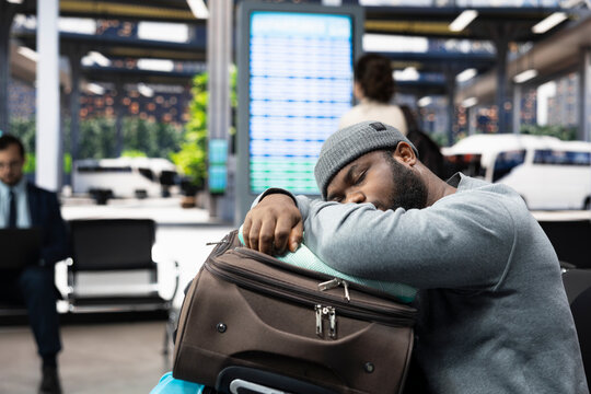 Migrant traveler dozes on stacked luggage in a coach station after carrying many bags through the terminal, resting on trolleys while worn out by job migration travel and a long wait.
