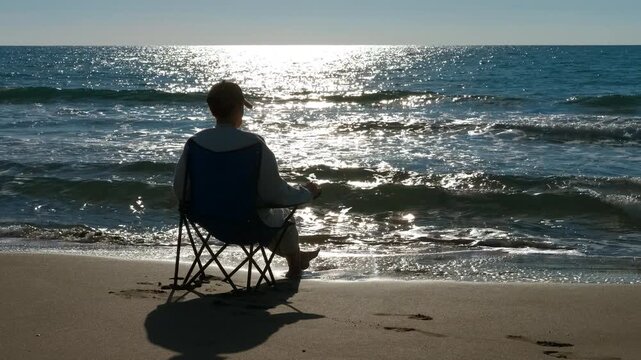 Person meditating sitting on a chair at the beach. Solitary person sitting on a folding chair at the seashore, meditating and watching the calm waves as the sun reflects on the ocean water