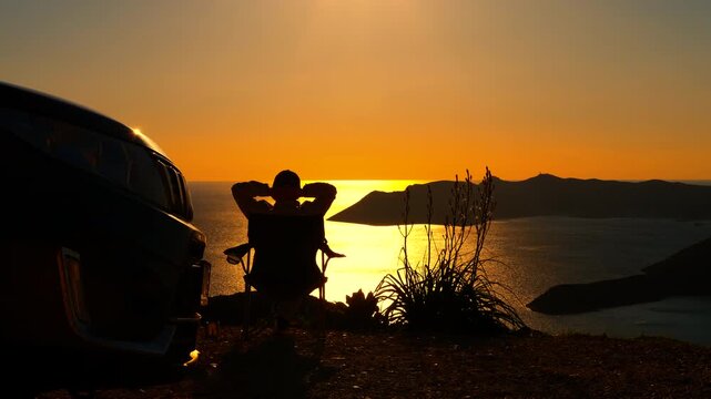Silhouette of a man relaxing watching the sunset over the sea. Silhouette of a relaxed traveler sitting on a camping chair next to his van, enjoying the stunning golden sunset view over the sea