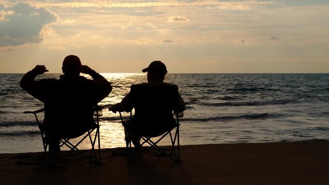 Two silhouetted men relaxing on beach chairs at sunset. Back view of two unrecognizable men sitting on folding chairs on a sandy beach, relaxing and enjoying the scenic view of the ocean