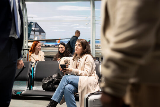 Woman sits on an airport chair holding a smartphone and dreaming of her travel destination, reminiscing about a finished holiday trip by checking vacation photos while waiting for a plane.