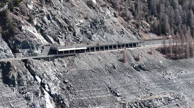 snow shed shields julier pass mountain road along crumbling scree slope waterfall spills down rocky gully sparse larch trees cling to steep mountainside aerial drone footage graubunden switzerland