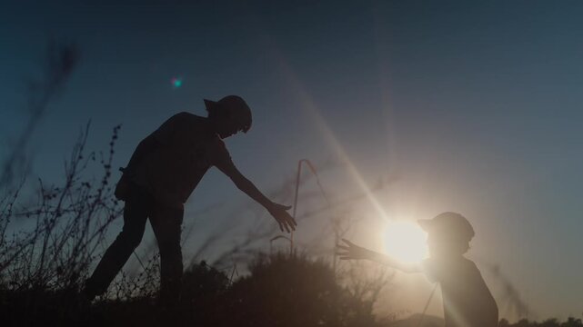 Silhouette of a boy helping his friend climb a hill at sunset, symbolizing childhood friendship, teamwork, and support in a beautiful natural landscape with lens flare