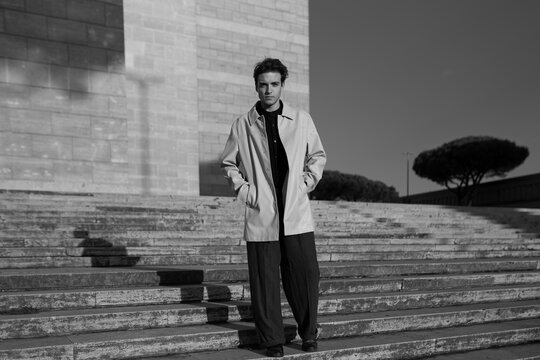 A young man in a stylish coat stands on wide stone steps under bright sunlight in black and white. Minimalist urban architecture frames quiet moments of modern elegance and personal confidence.