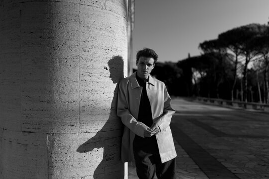 A young man in a coat leans against a stone column in a sunny outdoor setting in black and white. Timeless urban elegance inspires deep personal reflection and modern individual confidence.