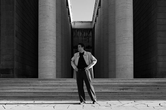 A young man in a stylish coat stands confidently before massive stone columns and wide stairs. Monumental urban architecture frames timeless modern elegance, personal ambition and deep authority.