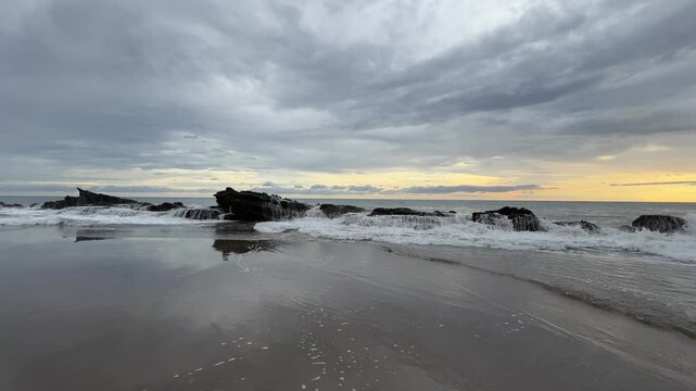 Waves crash over rocks at the beach as the sun sets behind clouds. The water reflects the sky and creates a serene view for all.