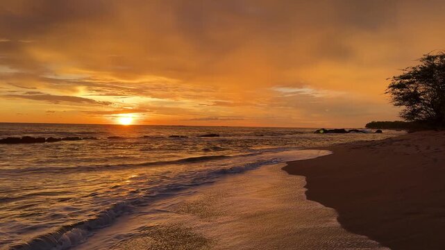 The sun sets over the ocean at a beach location. Waves gently crash on the sandy shore as colors fill the sky.