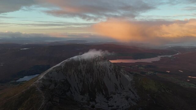 Wide drone view of snow dusted Errigal and lakes beneath glowing clouds, Donegal, Ireland