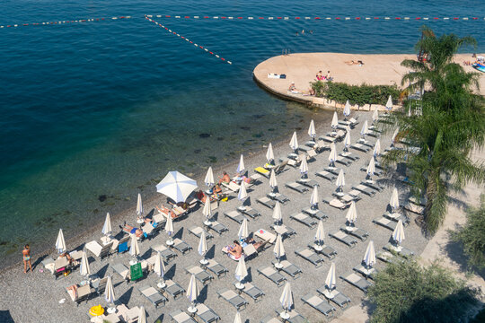 Tivat, Montenegro - 01 August 2025: View of a pebble beach in Tivat, Montenegro, with rows of sun loungers and white umbrellas along the clear blue Adriatic Sea.