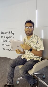 Corporate Laptop Using. A person wearing a patterned short-sleeve shirt and glasses while sitting in an office chair and holding a slim laptop.      