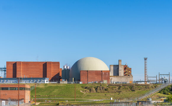 Nuclear power plant complex featuring a large dome containment structure, brick industrial buildings, power transmission lines, and green grassy hills