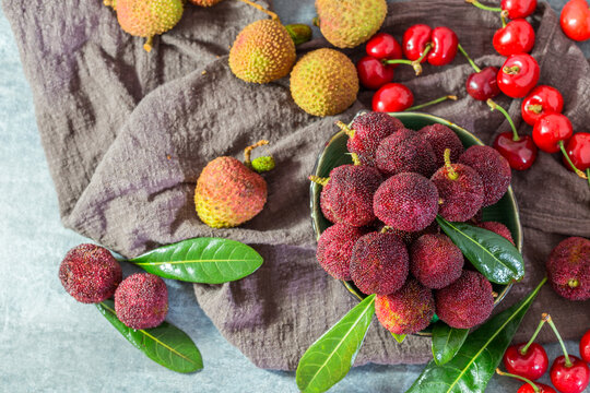 Fresh lychees and cherries are displayed in a bowl and on a textured cloth, accompanied by green leaves. This vibrant still life showcases ripe, colorful tropical and common fruits.
