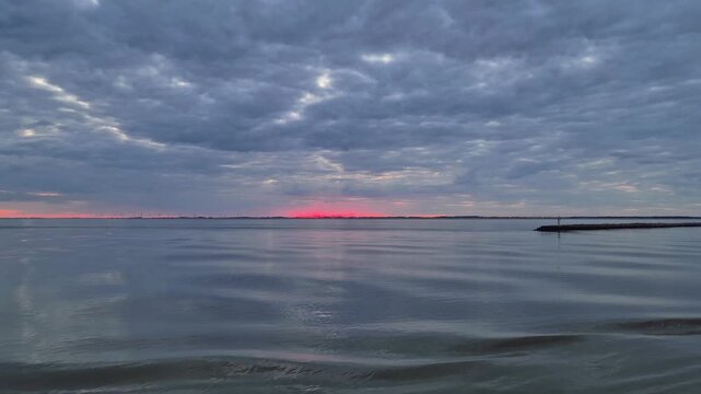 Scenic view of calm water ripples with vibrant red sunset glow at horizon under heavy cloudy overcast sky. Tranquil coastal seascape with a stone pier breakwater visible on the far right side