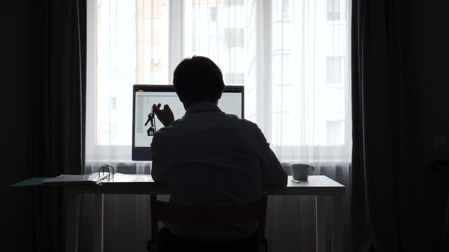 Person working from a dark home office, looking at a computer monitor while holding house keys, representing real estate, remote work, and property investment concepts
