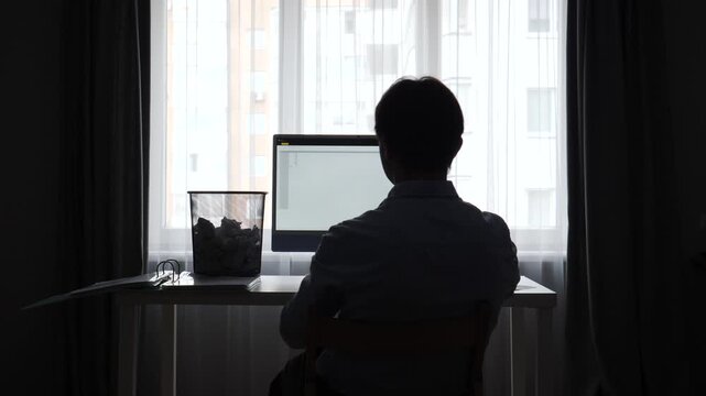 Man in silhouette experiencing frustration and stress while working from a home office, throwing crumpled paper into a wastebasket near a computer screen