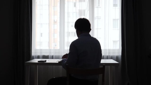 Man in silhouette holding an empty wallet, symbolizing financial crisis, debt, and poverty, sitting at a desk with pieces of paper on it in front of a bright window
