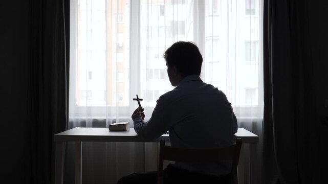 Man silhouetted at a table by a bright window, holding a small wooden cross and open bible in quiet reflection on faith, prayer, and spiritual solace