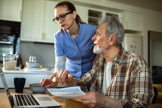 Caregiver helping senior man with paperwork on laptop at home